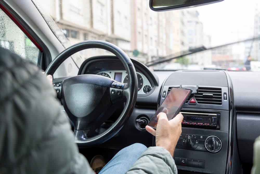 A male driver in a green jacket is dangerously using a smartphone while driving.
