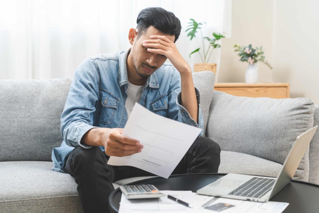 Stressed man reviewing bills and financial documents at home with laptop and calculator.